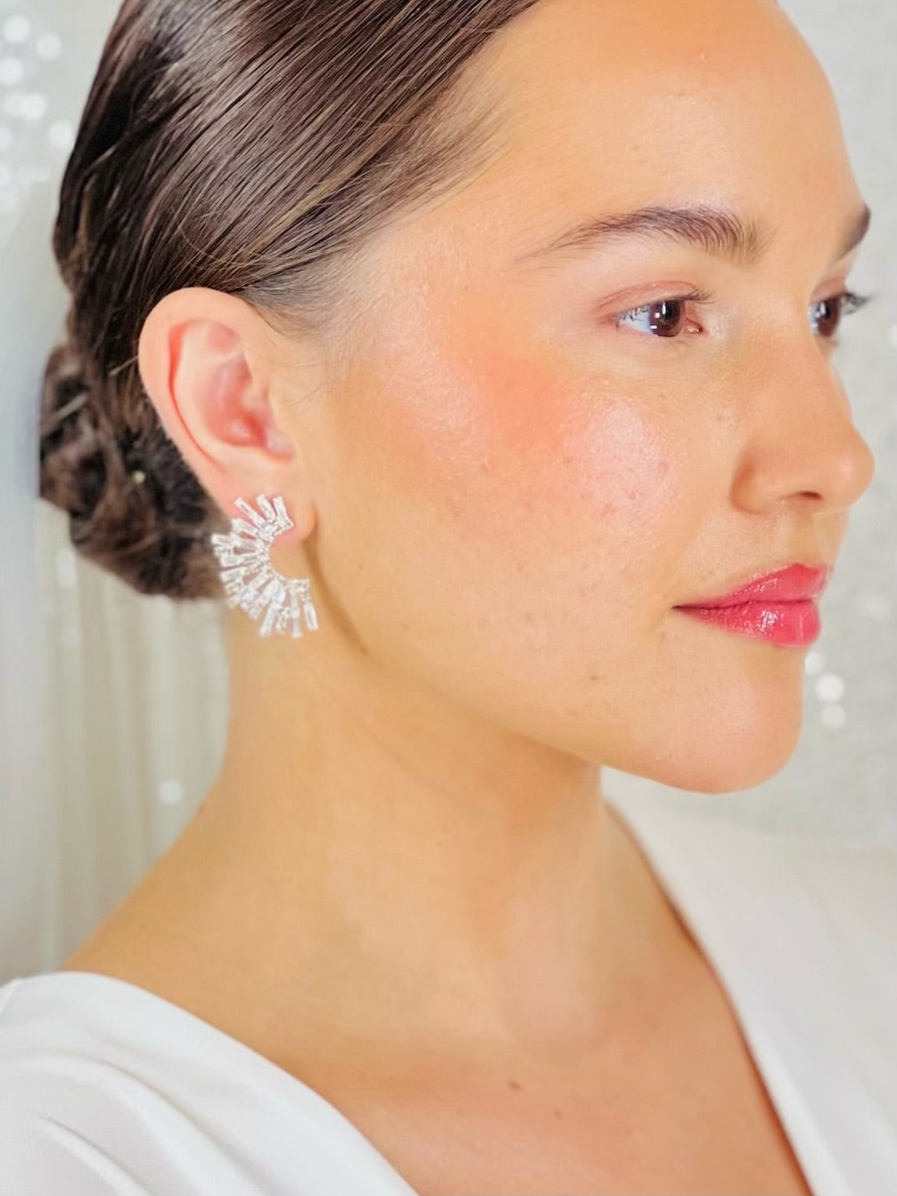 A woman with smooth, glowing skin and glossy pink lips wears her dark hair in a sleek bun, showcasing Cheveux Extensions by Mar's Silver Half Burst Earring—a stunning pick for bridal or elegant wedding jewelry.